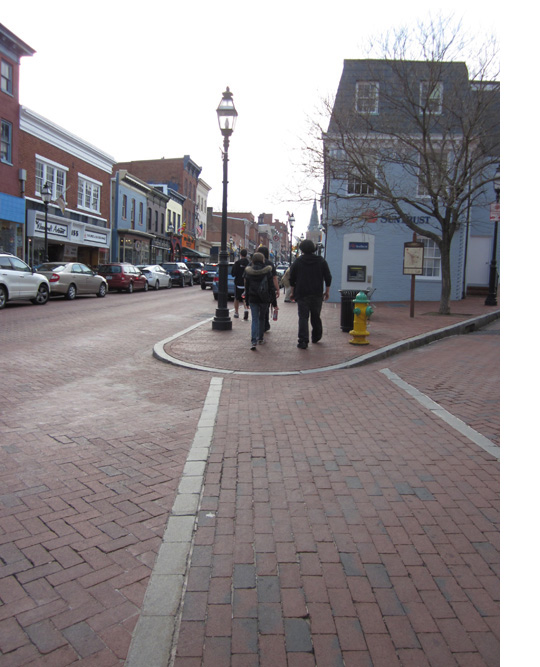 Photo shows two brick streets intersecting at an angle of about 30 degrees, forming a very acute corner.  The sidewalk is made out of brick, and slopes gently down to the street at the apex of the corner, with about 4 feet of the edge of the sidewalk being flush with the street.